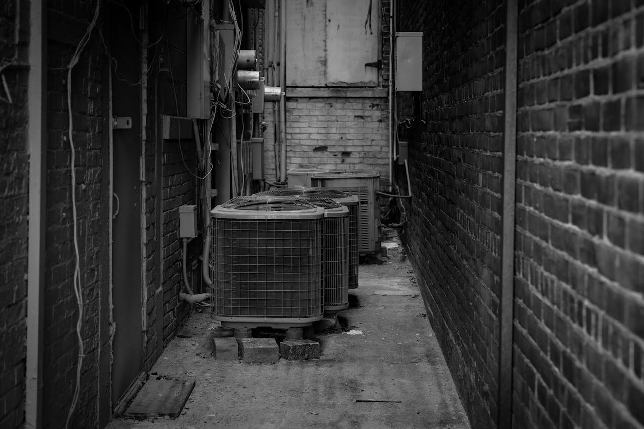 Black and white alley featuring industrial air conditioning units against a brick wall.